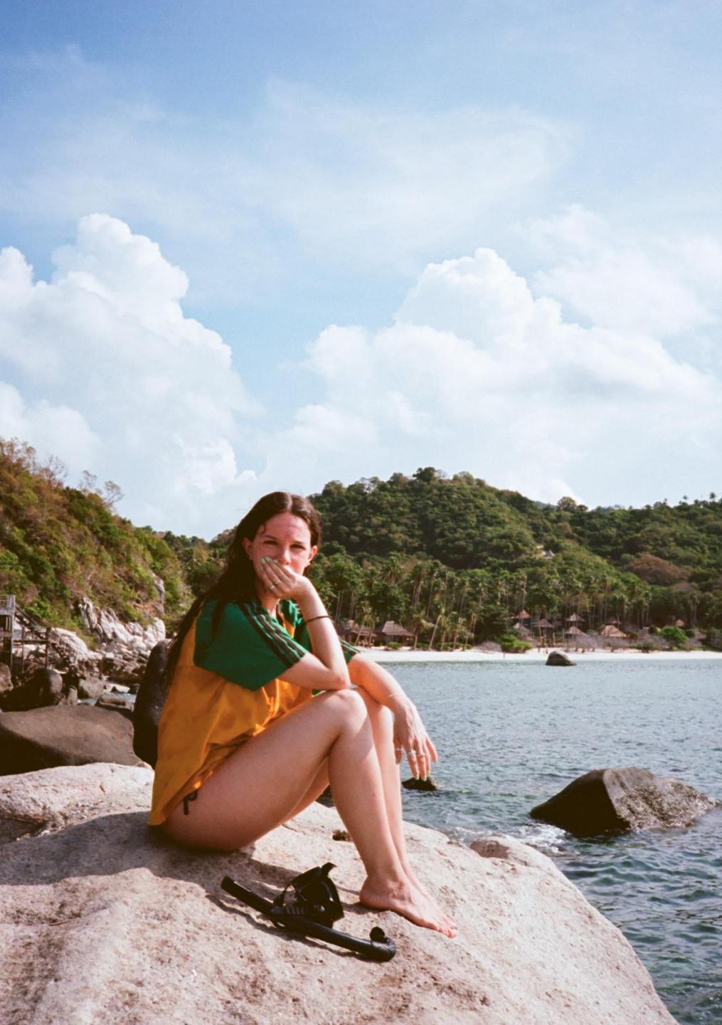 A woman sits on a large rock by the ocean with a tropical island in the background.