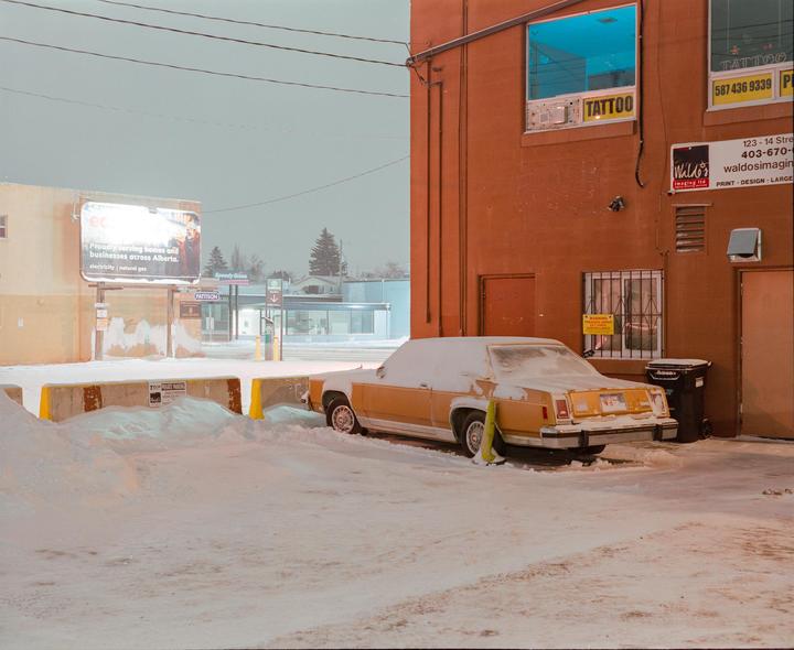 A snow-covered yellow car is parked next to a brick building with a tattoo sign.