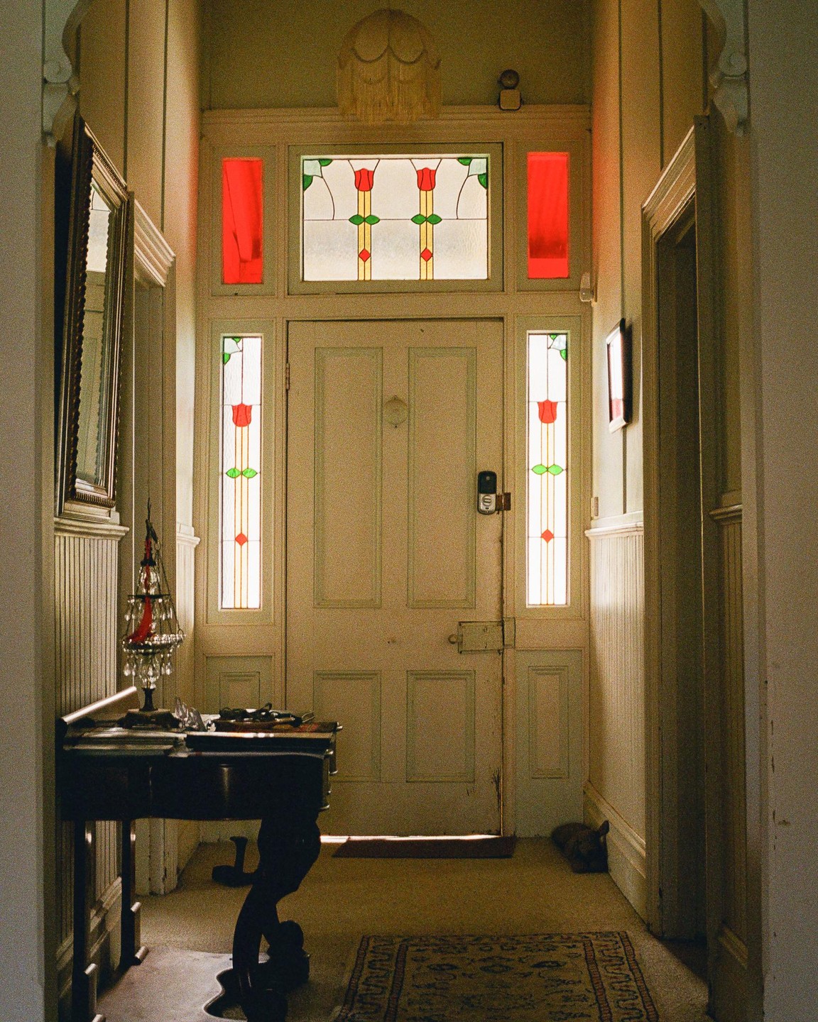 Sunlight streams through a front door with stained glass panels, illuminating a hallway with a console table and a small rug.