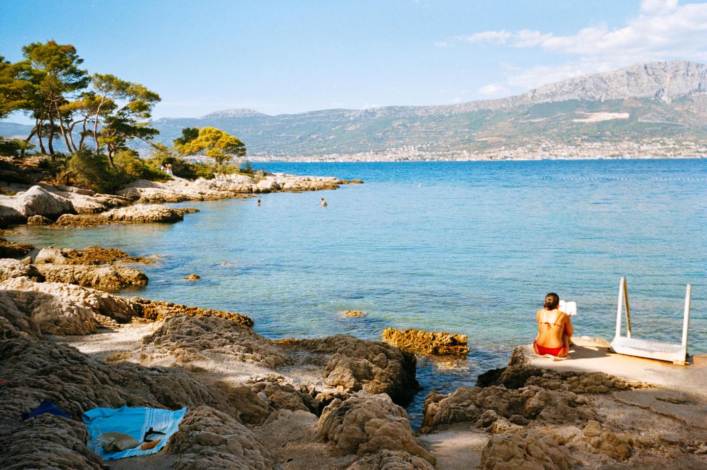 A woman sits on a concrete slab by the sea, with mountains in the distance.