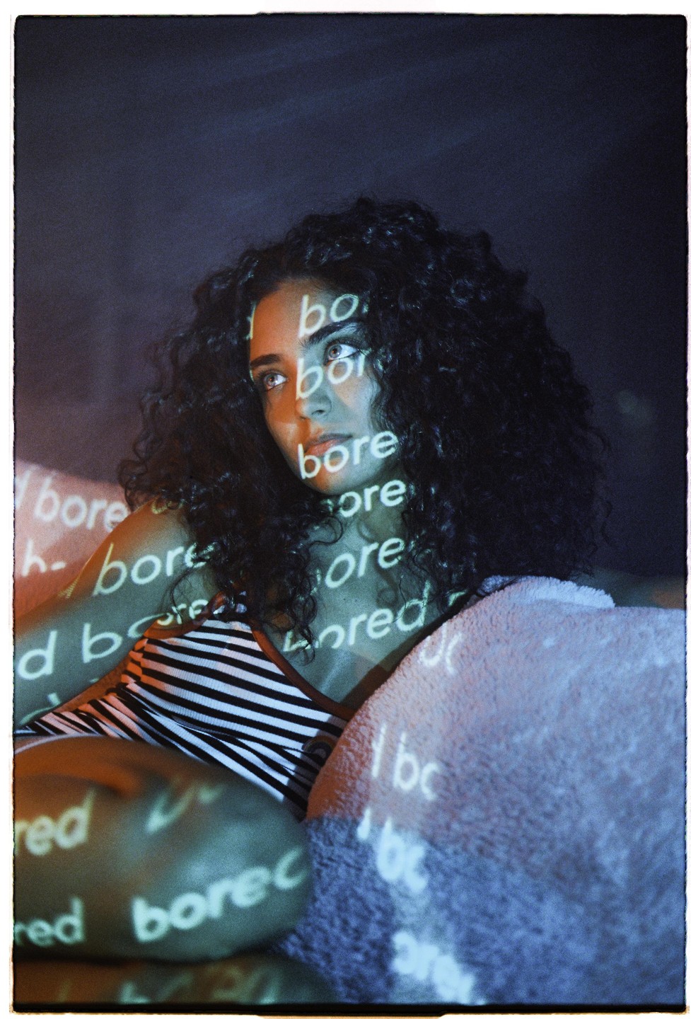 A woman with curly hair is illuminated by projected text that reads "bore" and "bored" across her face and body.
