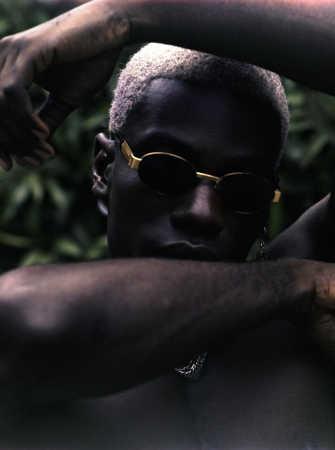 A man with bleached hair and sunglasses poses with his arms crossed over his head and chest.