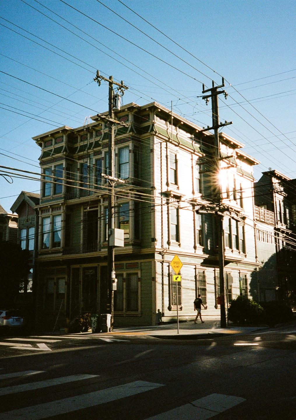 A large, ornate building with many windows stands on a street corner under a clear sky, with power lines crisscrossing above.