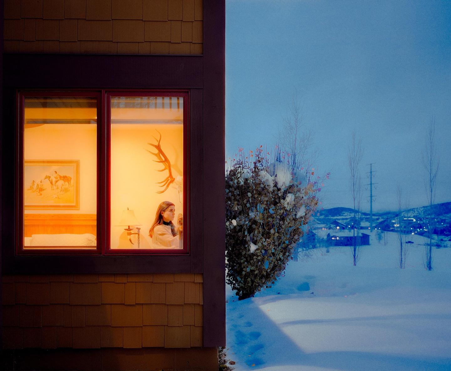 A woman looks out a window from a warmly lit room into a snowy landscape at dusk.