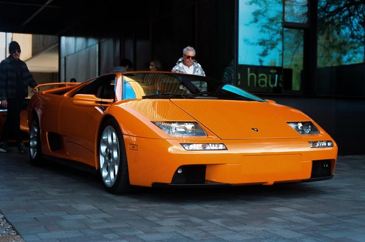 An orange sports car is parked on a paved surface with people standing in the background.