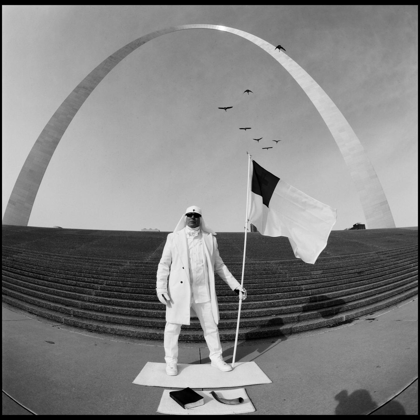 A man in white attire holds a flag while standing in front of the Gateway Arch.