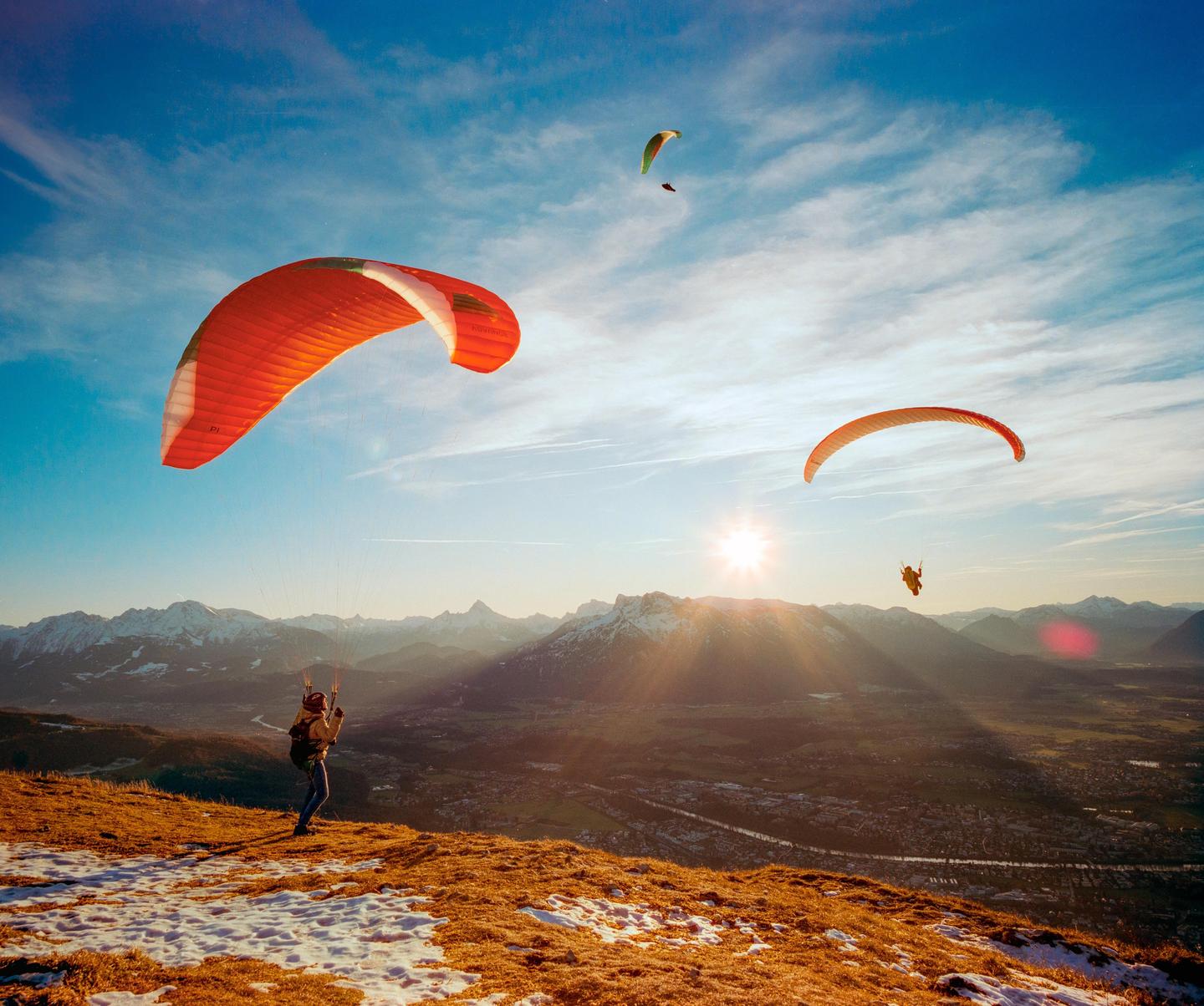 A person prepares to paraglide from a snowy mountain peak overlooking a valley with other paragliders in the sky.