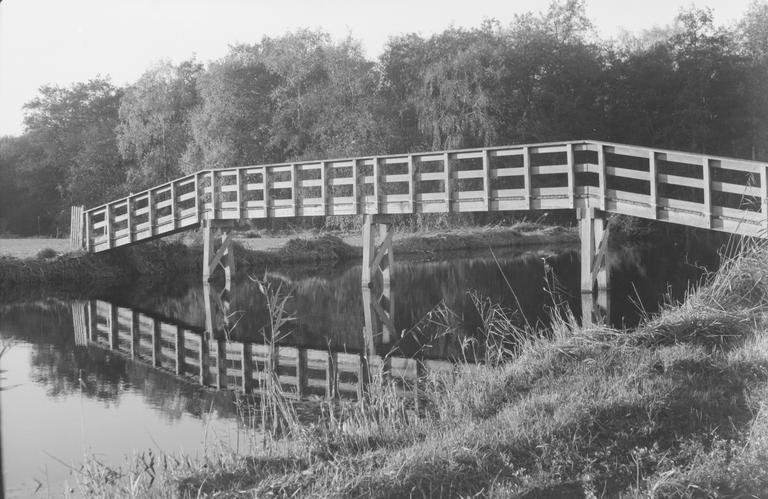 A wooden footbridge crosses a narrow body of water, reflecting in the still surface.