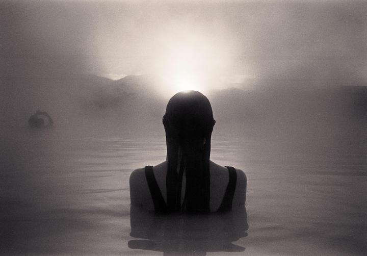 A woman with long hair is seen from behind, submerged in water with a bright light source on the horizon.