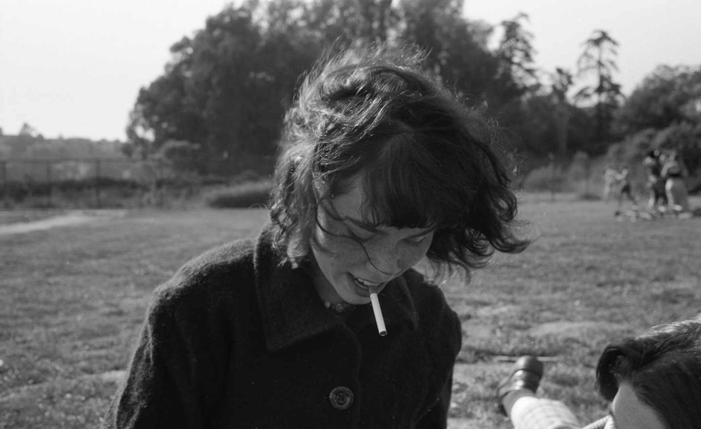 A woman with windblown hair and freckles smiles with a cigarette in her mouth in a grassy field.