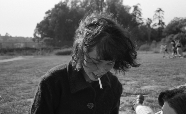 A woman with windblown hair and freckles smiles with a cigarette in her mouth in a grassy field.