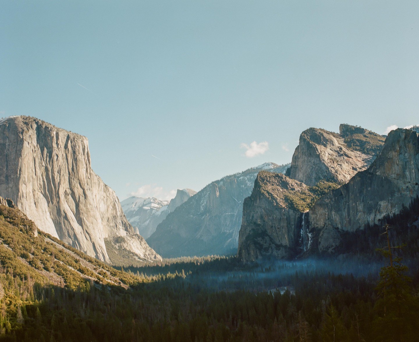 A valley with a forest and a waterfall is surrounded by mountains under a clear sky.