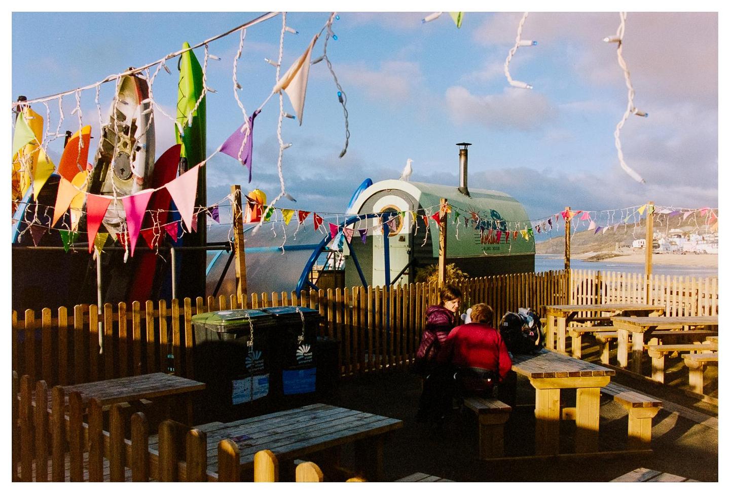 Two women sit at a picnic table in an outdoor eating area decorated with colorful bunting and string lights, with a beach and ocean visible in the background.
