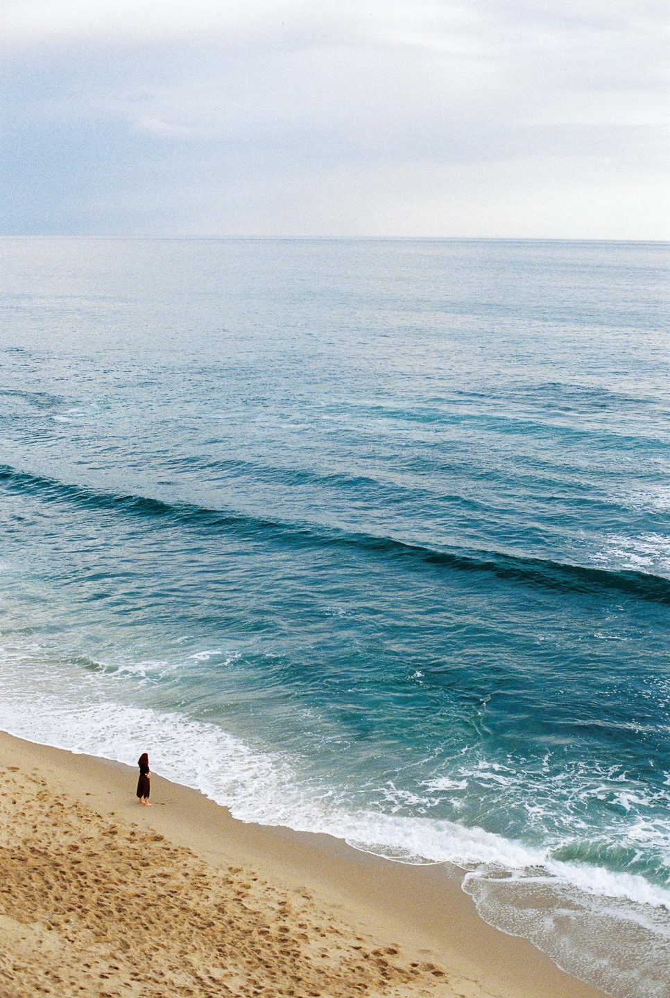 A woman stands on a sandy beach looking out at the ocean with gentle waves.