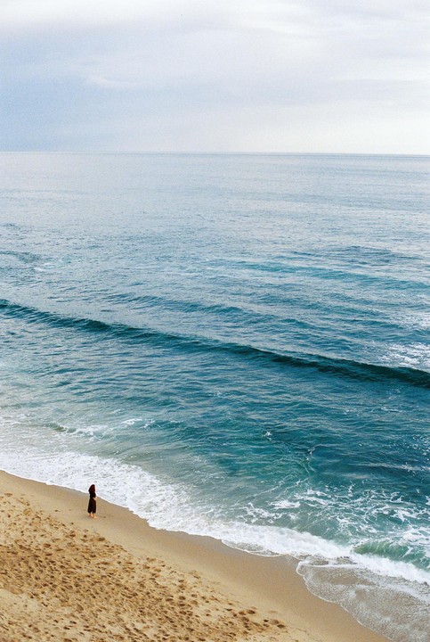 A woman stands on a sandy beach looking out at the ocean with gentle waves.