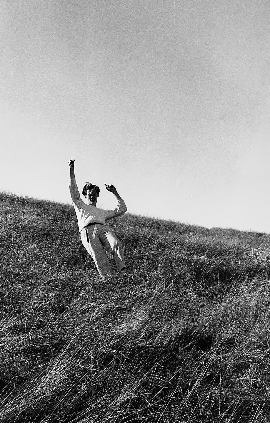 A man in light clothing is captured mid-fall on a grassy hillside under a bright sky.