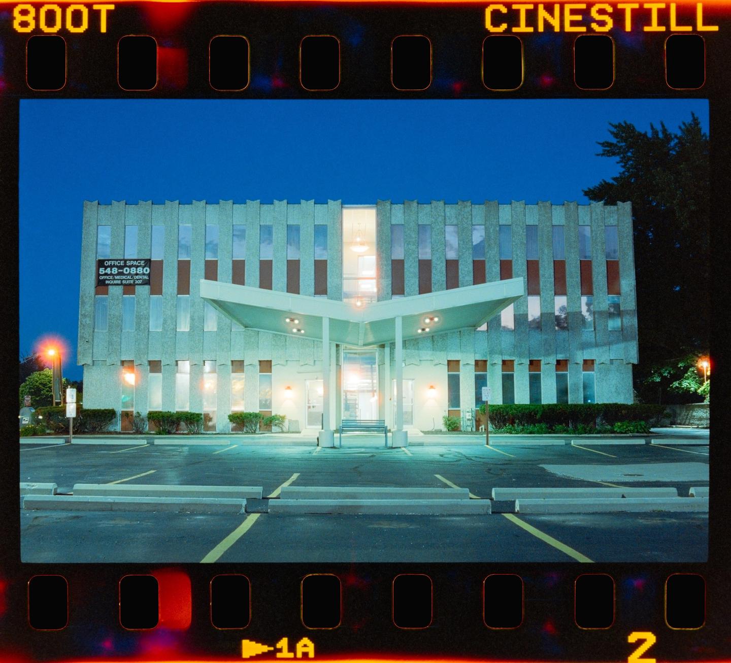 A building with a large awning over its entrance stands next to a parking lot at night.