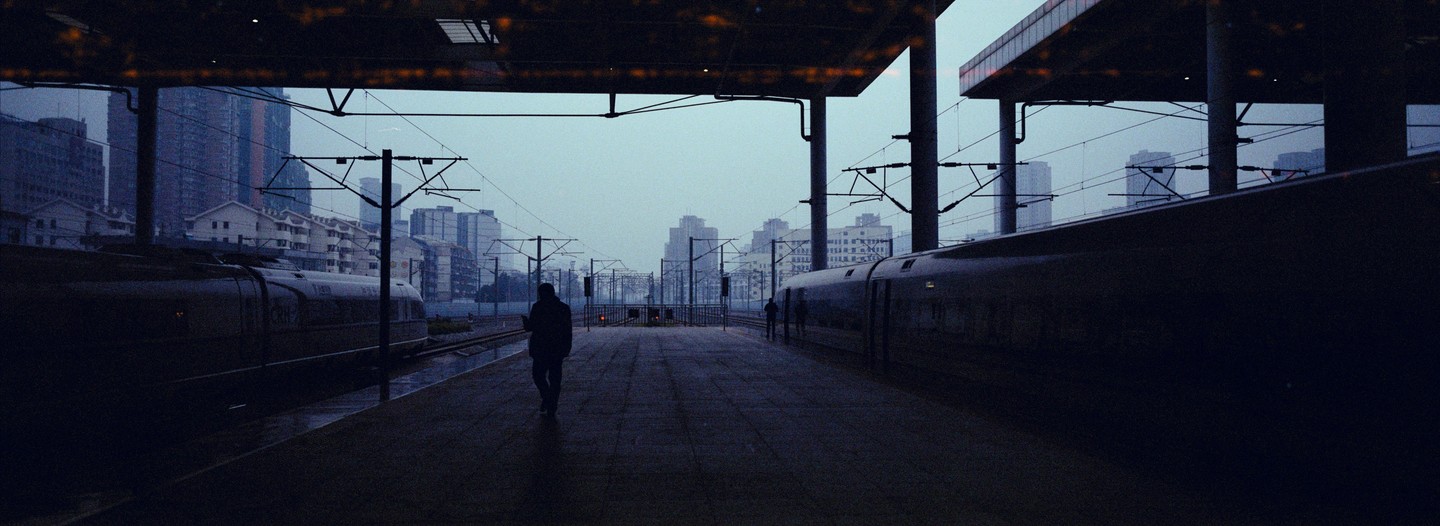 A man walks along a train platform with two trains on either side and a city skyline in the background.
