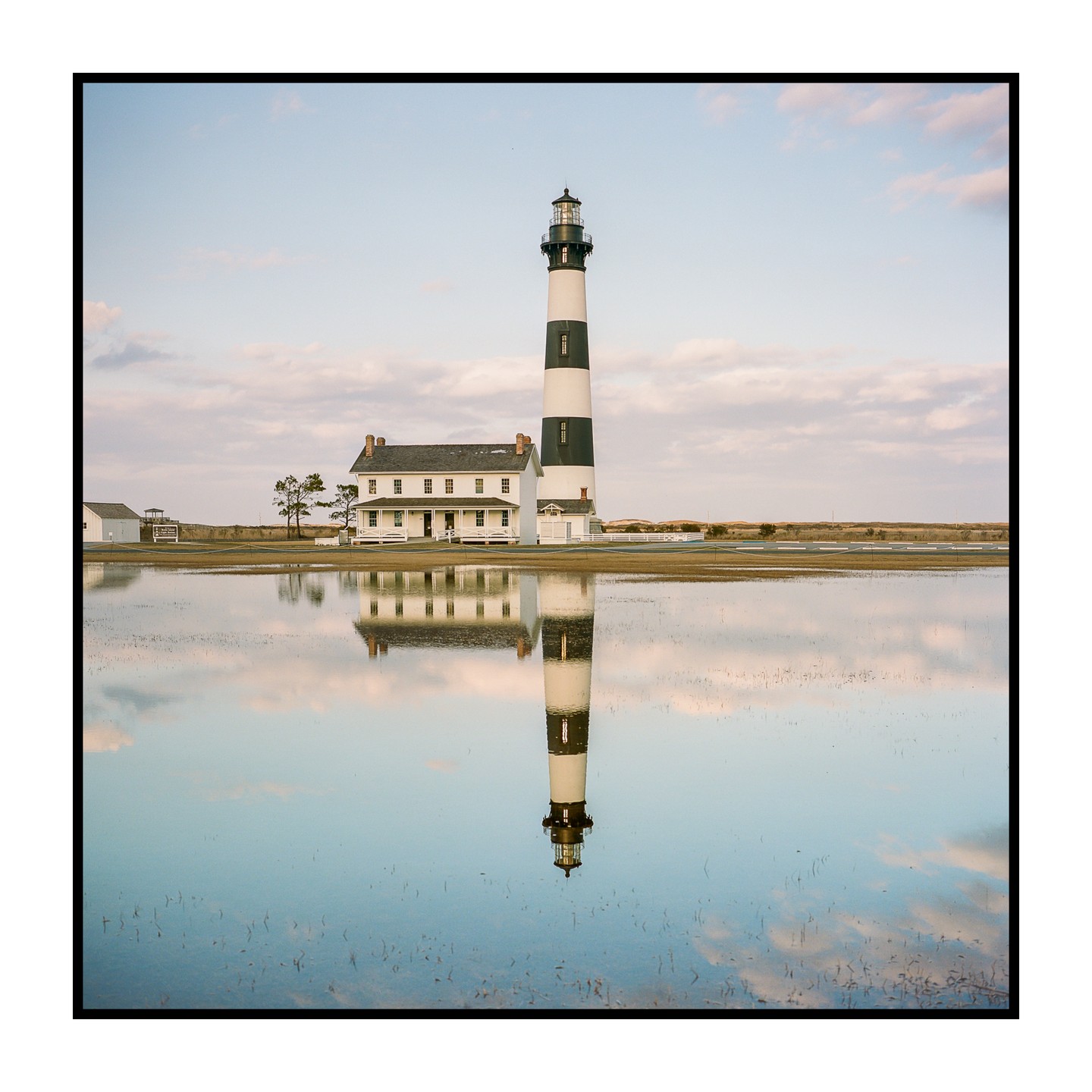 A tall, striped lighthouse stands next to a white building, both reflected in a still body of water under a cloudy sky.