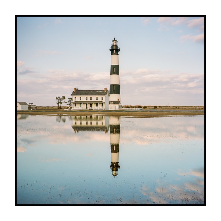A tall, striped lighthouse stands next to a white building, both reflected in a still body of water under a cloudy sky.