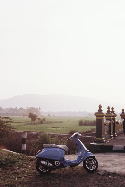 A blue scooter is parked on a dirt road next to a bridge with a rice field and mountains in the background.