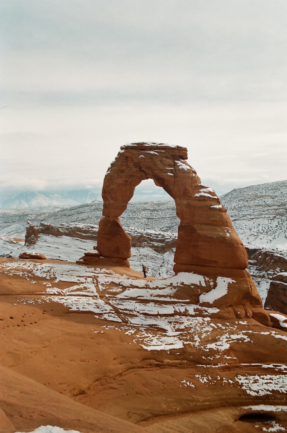A person stands under a large natural arch in a snowy, mountainous landscape.