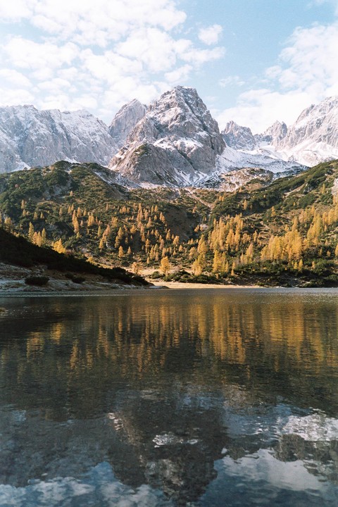 A lake reflects snow-capped mountains and a forest of golden trees under a cloudy sky.
