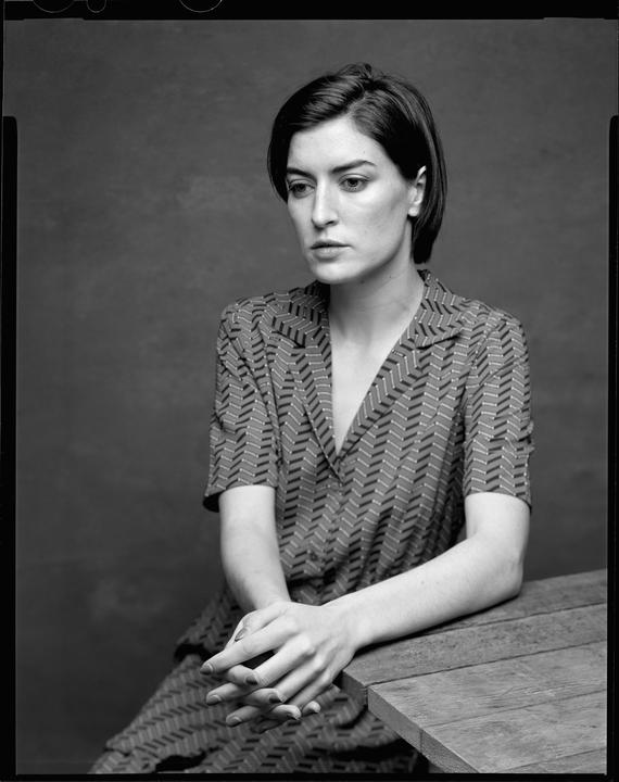 A woman with short hair and a patterned shirt sits at a wooden table with her hands clasped.