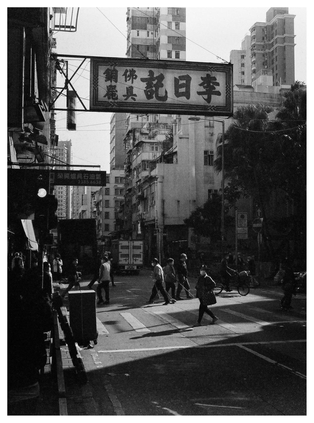 A street scene in Hong Kong shows people crossing a crosswalk under a large sign with Chinese characters.