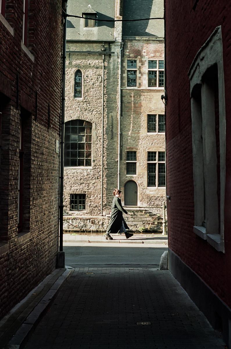 A woman in a long coat walks along a narrow street between brick buildings.