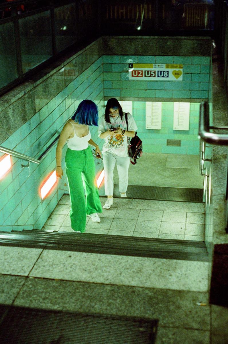 Two women descend a subway staircase with teal tiled walls and orange-lit handrails.