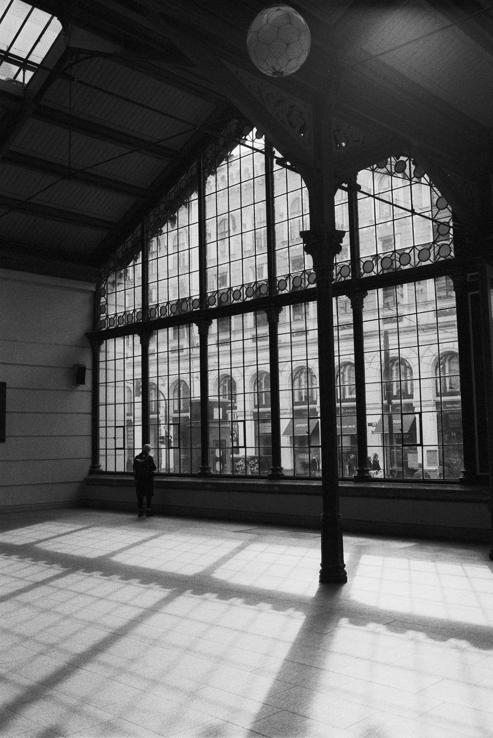 A person stands in a large hall with a tall, arched window looking out onto a building across the street, with sunlight casting grid shadows on the floor.
