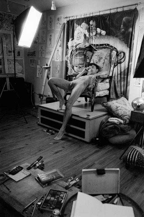 A nude man reclines in an ornate chair in an artist's studio.