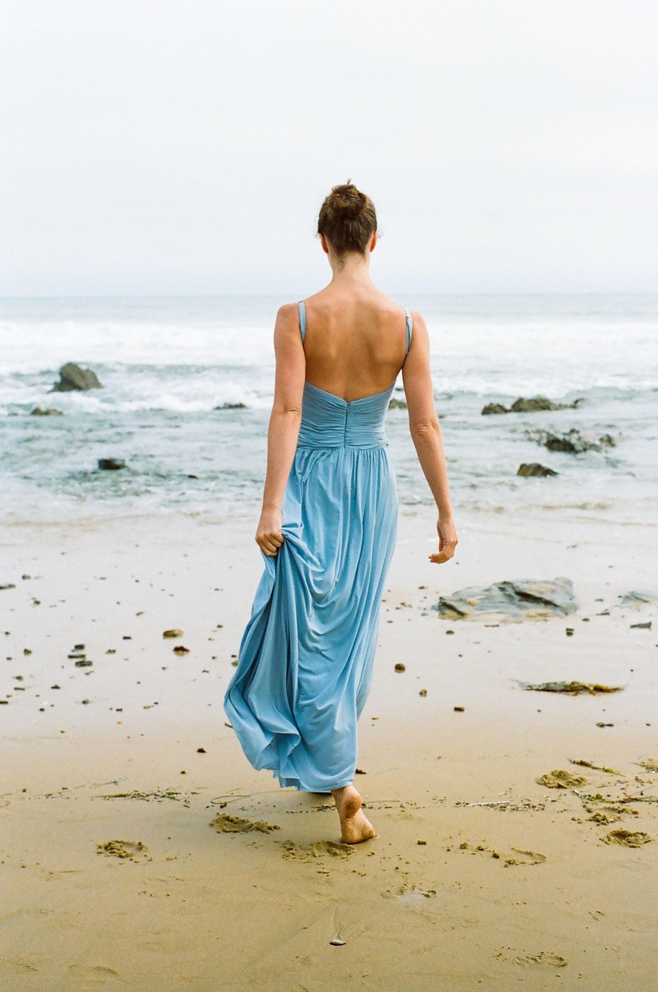 A woman in a blue dress walks barefoot on a sandy beach towards the ocean.