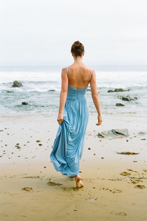 A woman in a blue dress walks barefoot on a sandy beach towards the ocean.