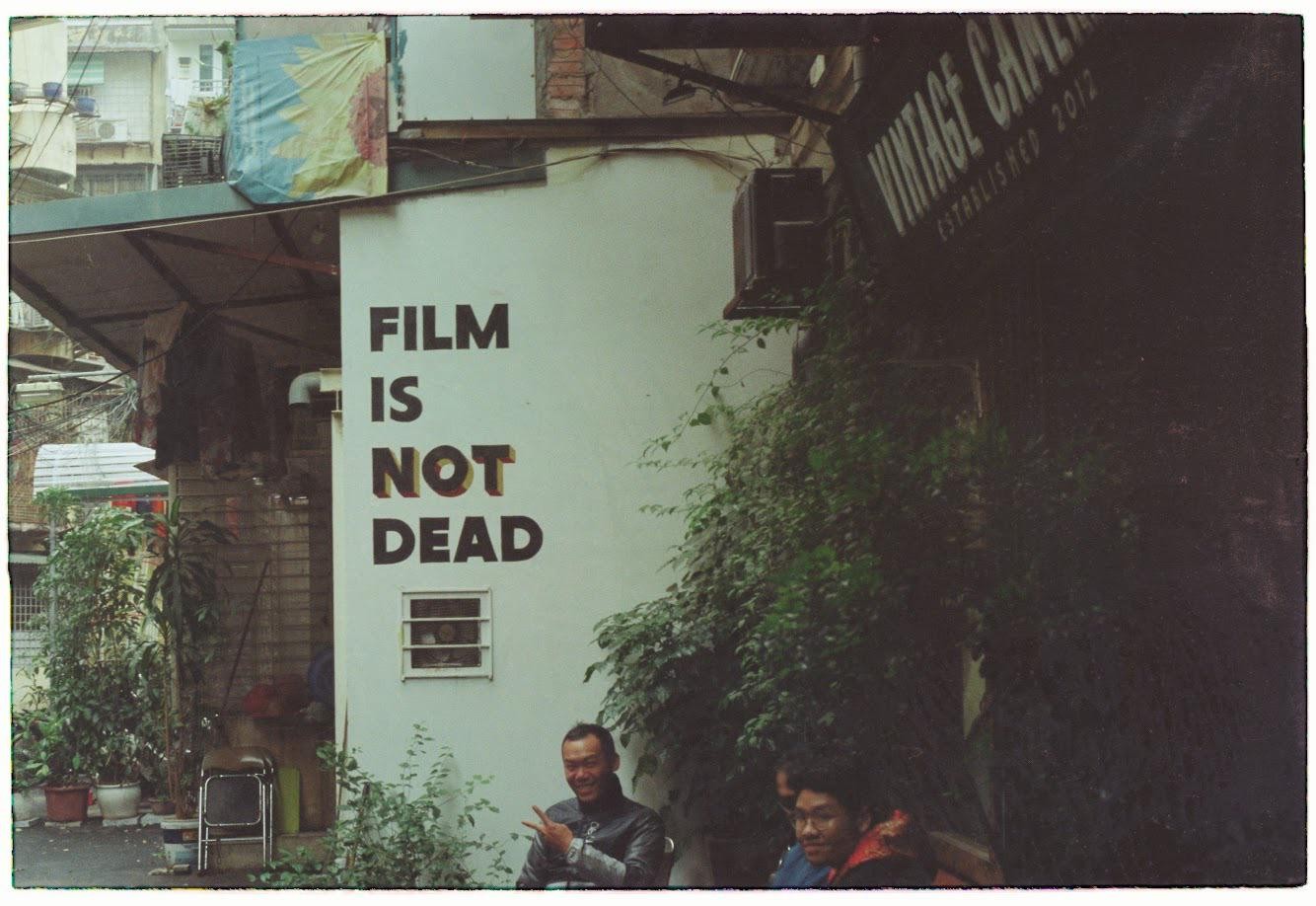 A man makes a peace sign in front of a wall with "FILM IS NOT DEAD" painted on it, next to a vintage camera store.