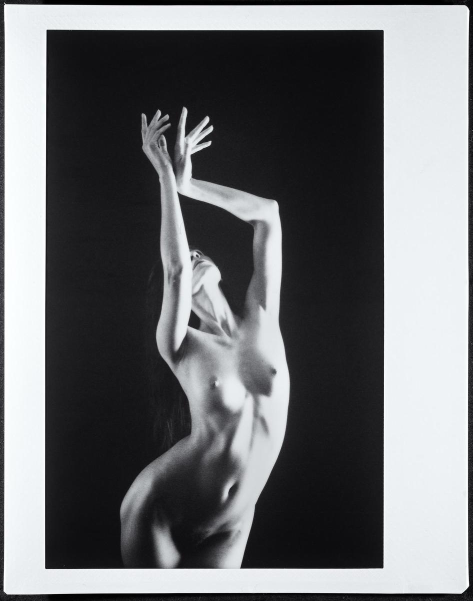 A nude woman with her arms raised and head tilted back is captured in a studio setting.