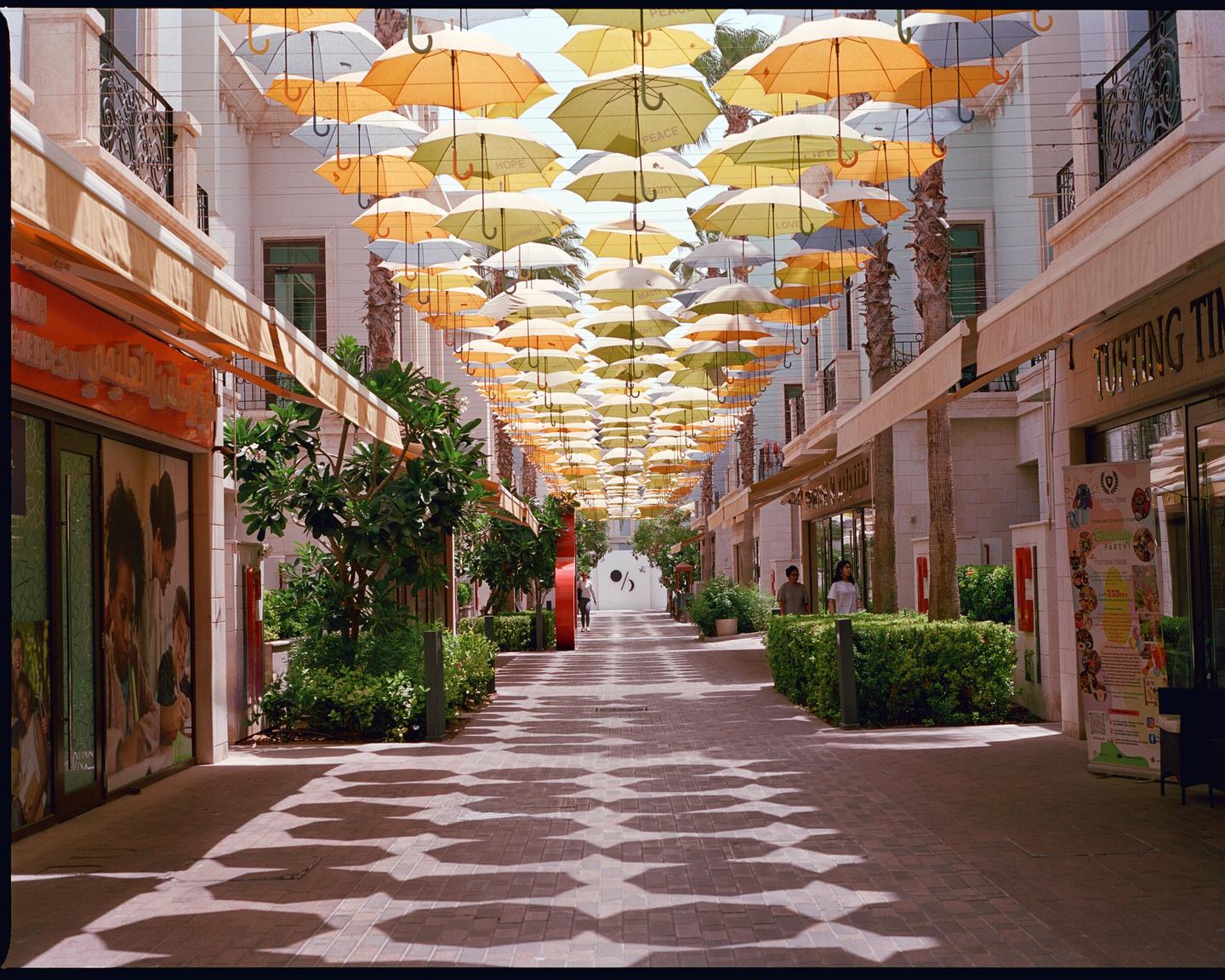 A pedestrian street is decorated with many yellow and white umbrellas hanging overhead, casting shadows on the ground.