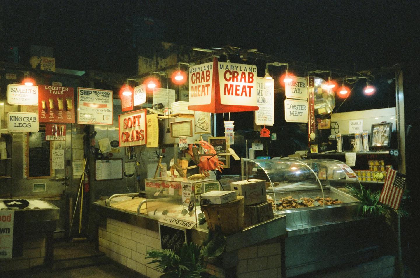 A seafood market at night is illuminated by red lights and signs advertising crab meat, lobster, and oysters.