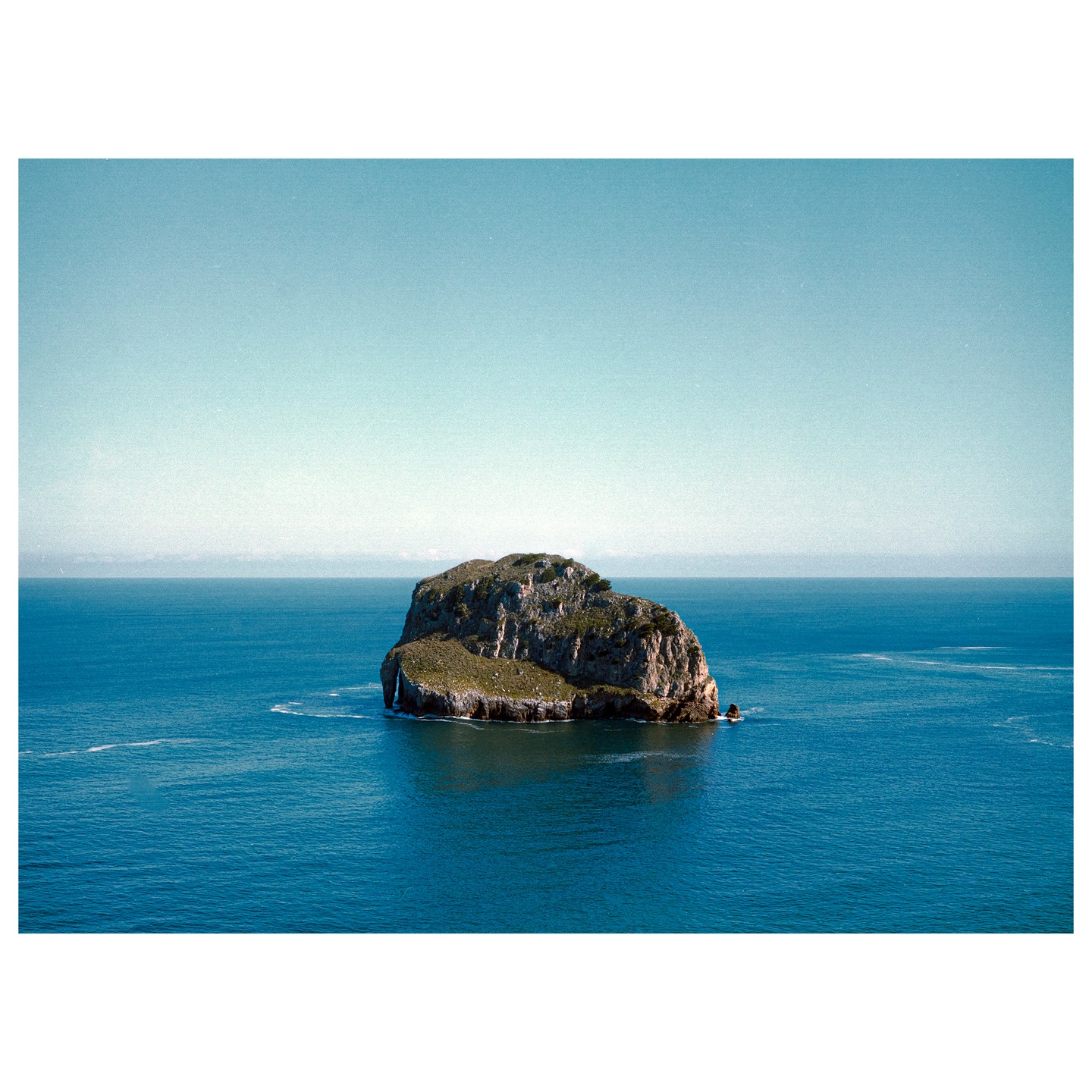 A small, rocky island covered in green vegetation sits in the middle of a vast blue ocean under a clear sky.