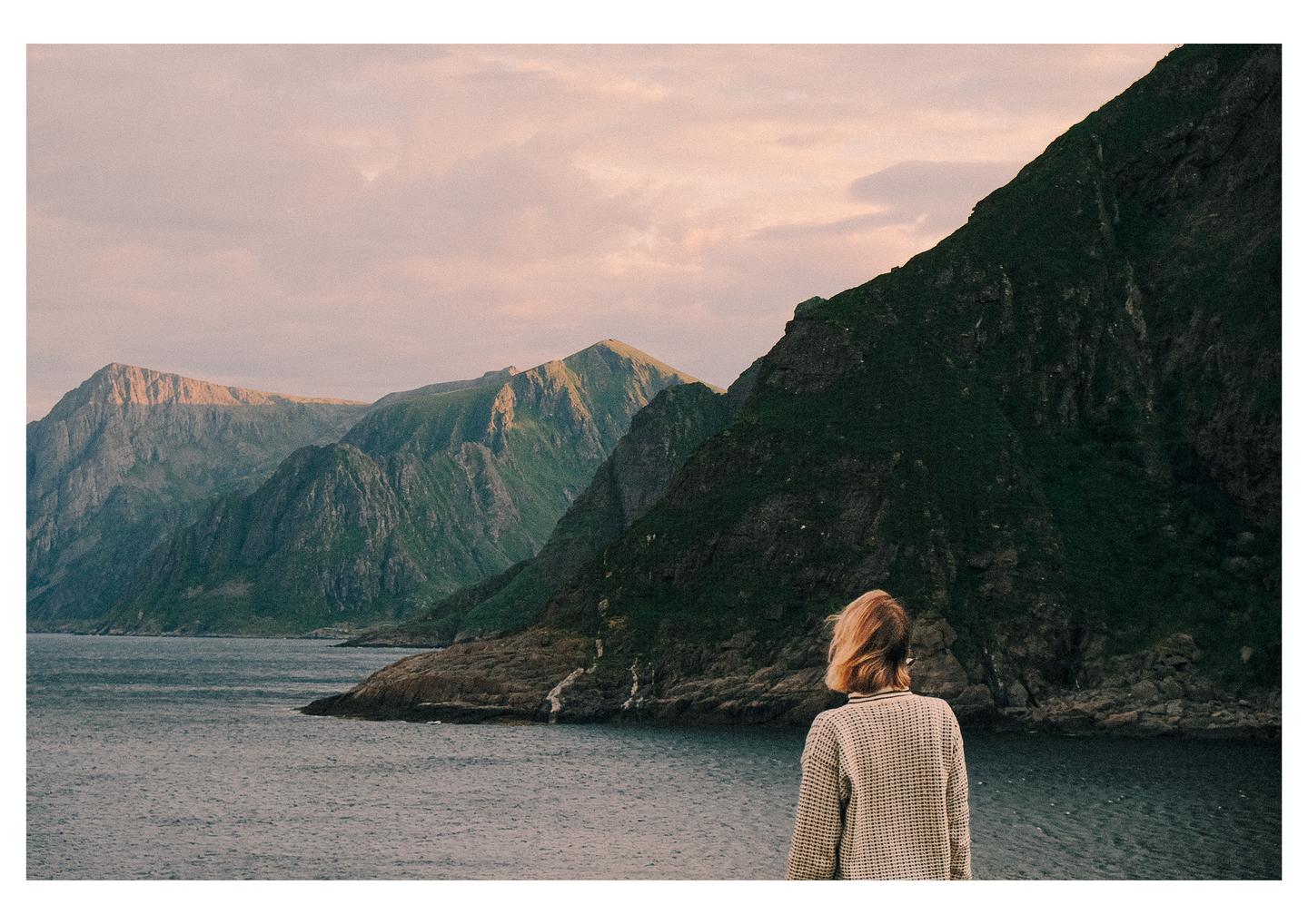 A woman stands with her back to the viewer, looking out at a body of water with mountains in the distance under a cloudy sky.