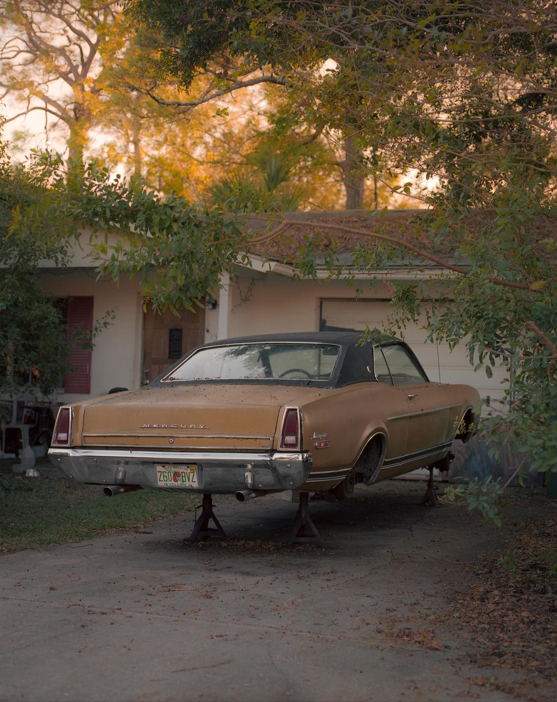 A gold vintage car is parked on stilts in a driveway in front of a house.
