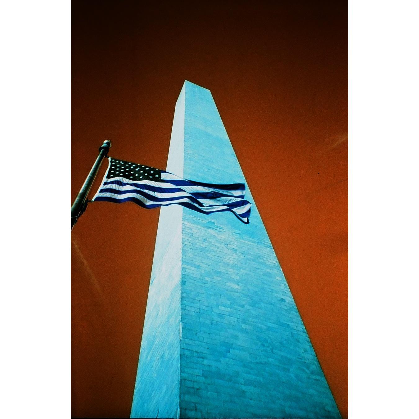 An American flag flies in front of the Washington Monument against an orange sky.