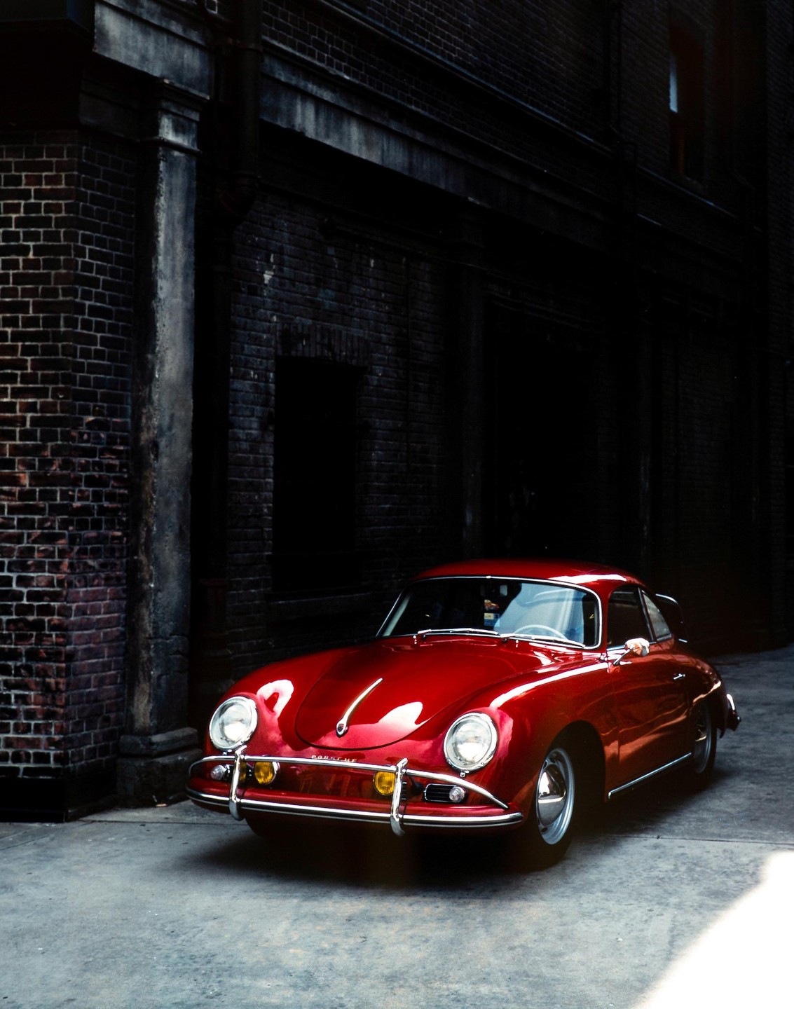 A cherry red vintage car is parked in a dark alleyway next to a brick building.