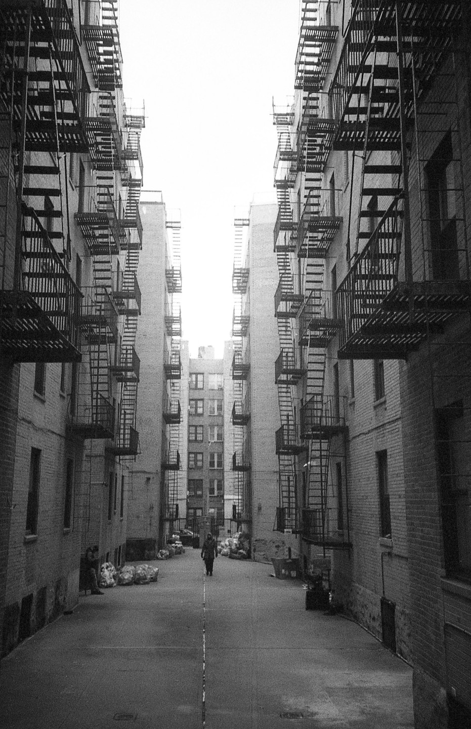 A person walks down a narrow alleyway between two tall buildings with fire escapes in Harlem, New York.