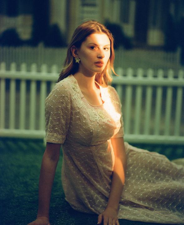 A woman in a light pink dress sits on grass in front of a white picket fence.