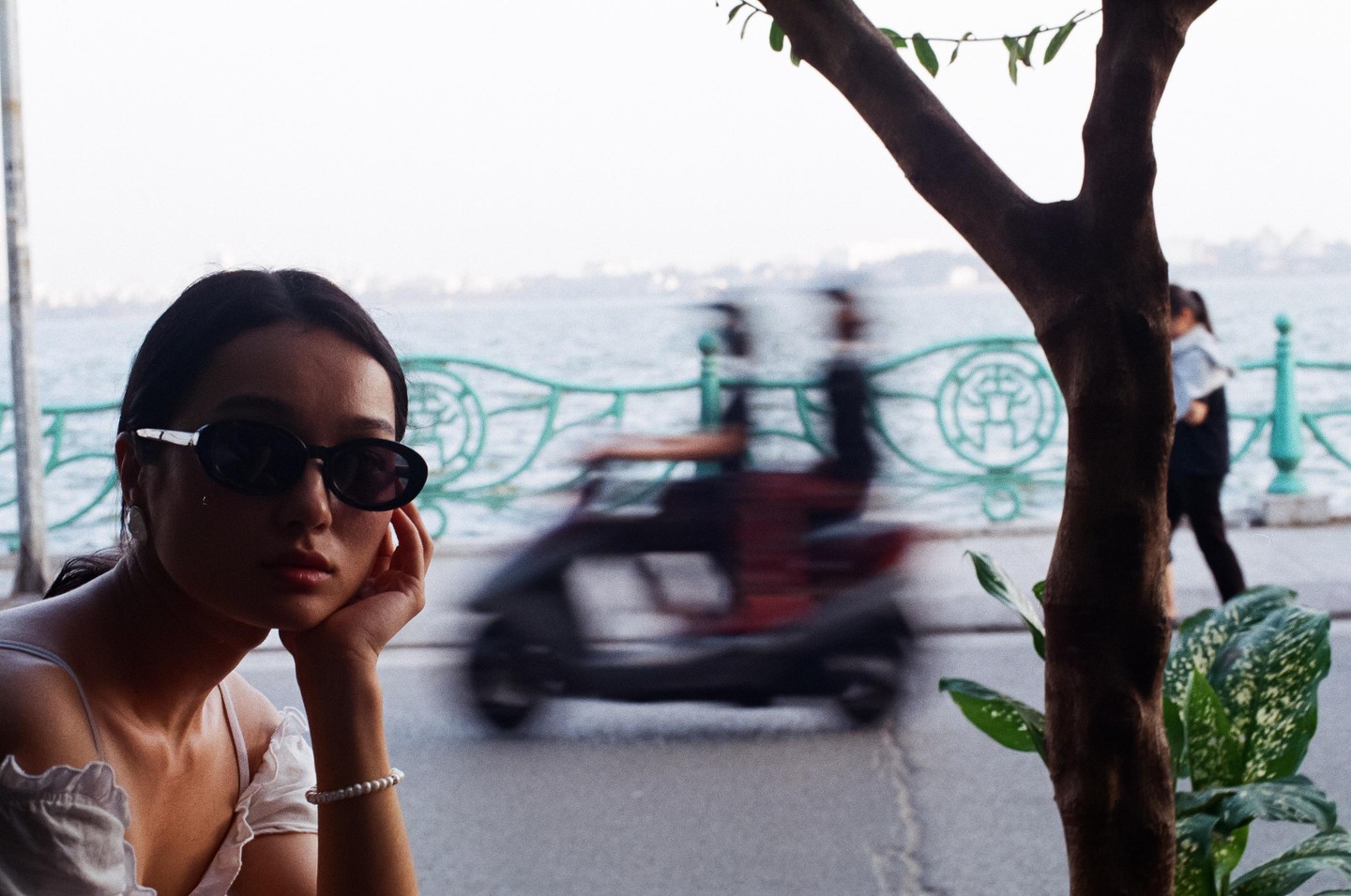 A woman in sunglasses rests her chin on her hand, with a blurred scooter and lake in the background.