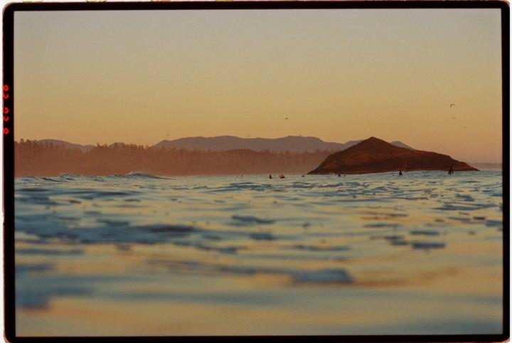 A body of water with small waves is in the foreground, with a small island and mountains in the background under a golden sky.