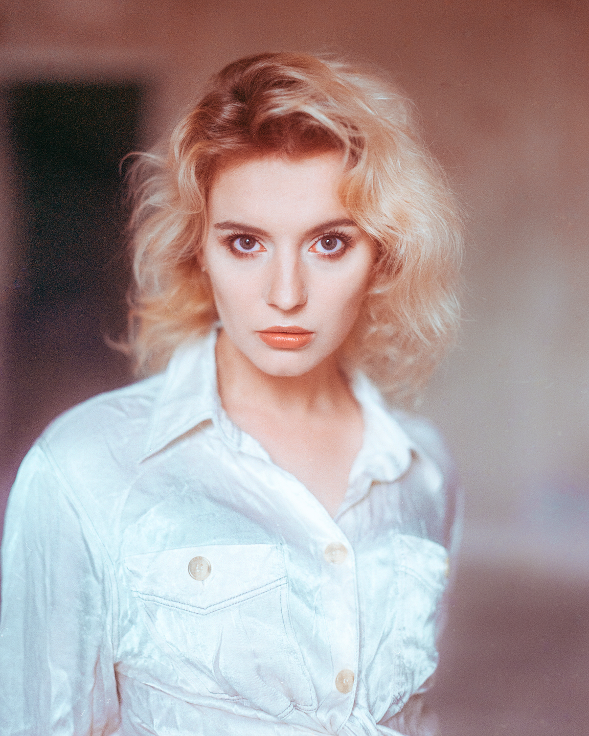 A woman with curly blonde hair and a white shirt looks directly at the viewer.