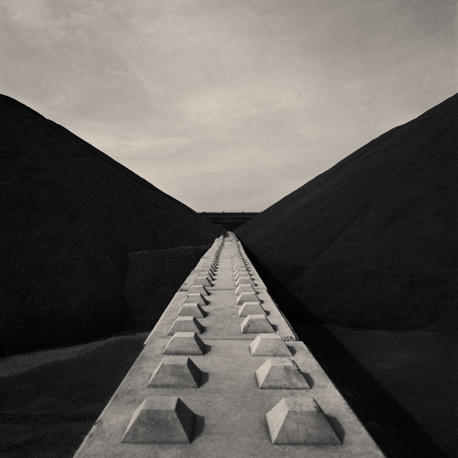 A concrete barrier with pyramid-shaped protrusions extends into the distance between two large, dark mounds under a bright sky.